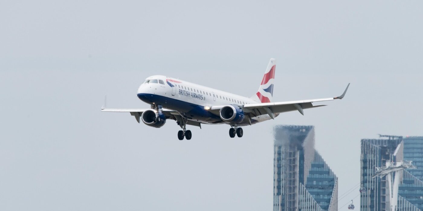 A British Airways plane is taking off against a grey sky. Some London skyscrapers can be seen in the background.