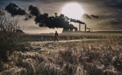 A person walking across a field with thick smoke coming out of a factory in the background / Image: Unsplash