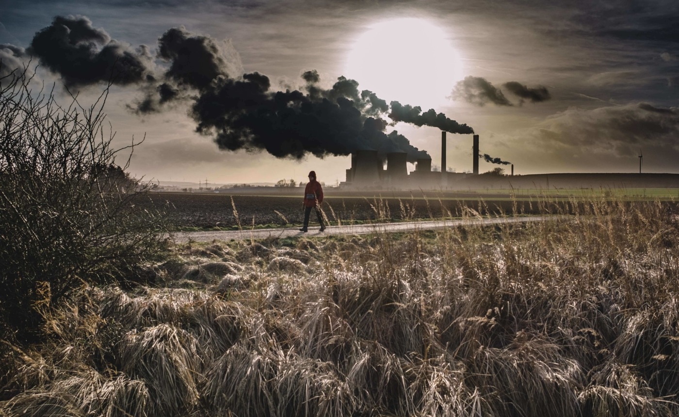A person walking across a field with thick smoke coming out of a factory in the background / Image: Unsplash