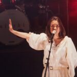 Singer Mitski performs on stage. She is wearing a white dress and has her right arm extended to the side