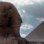 A sphinx in focus in the foreground and the blurry Pyramid of Giza in the background, against a dramatically cloudy sky.