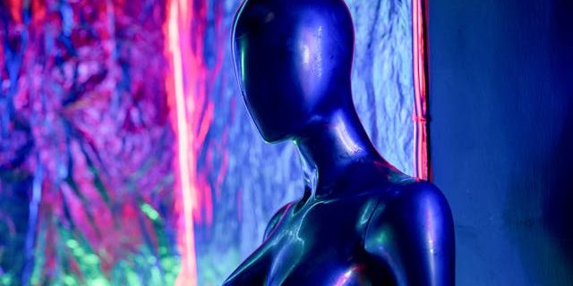 Side view of a black mannequin with purple, red, blue and green light behind it. Some reflection of purple light on the mannequin itself.