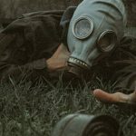 Image shows a man lying on the ground wearing a gas mask. He is holding his neck and reaching for a can with the other.