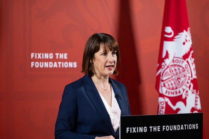 UK Chancellor Rachel Reeves giving a speech. Text saying "Fixing the Foundations" is enscribed on her lectern and the red background behind her.