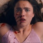 Close-up of a distressed, young, white, brunette female lying on her back on a cream carpet with splatters of blood across her face and upper body