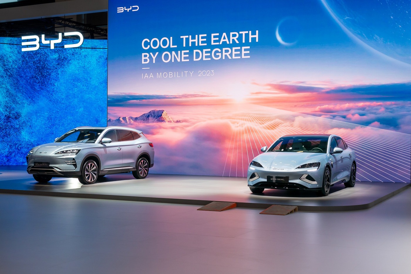 Two BYD electric cars at a car show, one SUV and one sedan, in front of a background photo of the sky.