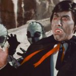 A man in a humorous vampire costume holds his hands up in dismay with two costume aliens behind him