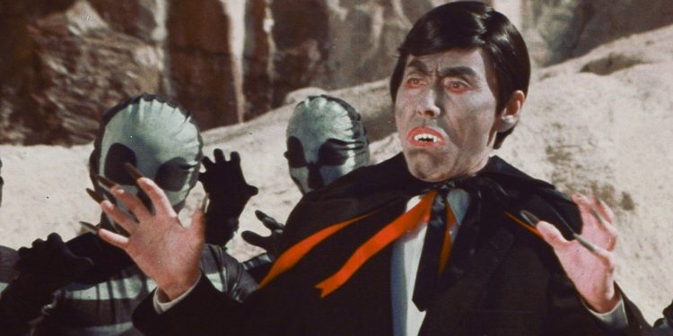 A man in a humorous vampire costume holds his hands up in dismay with two costume aliens behind him