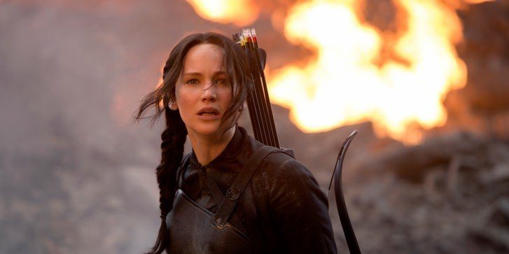 A brunette white woman looks shocked and scared with a blaze of fire behind her as she carries a bow and arrow on her back
