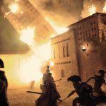 Ancient greek soldiers in a scene of the fall of Troy fight within the city as a burning building falls behind them