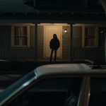 A car is parked outside of an American suburban home at night and a woman's silhouette stands in the porchlight