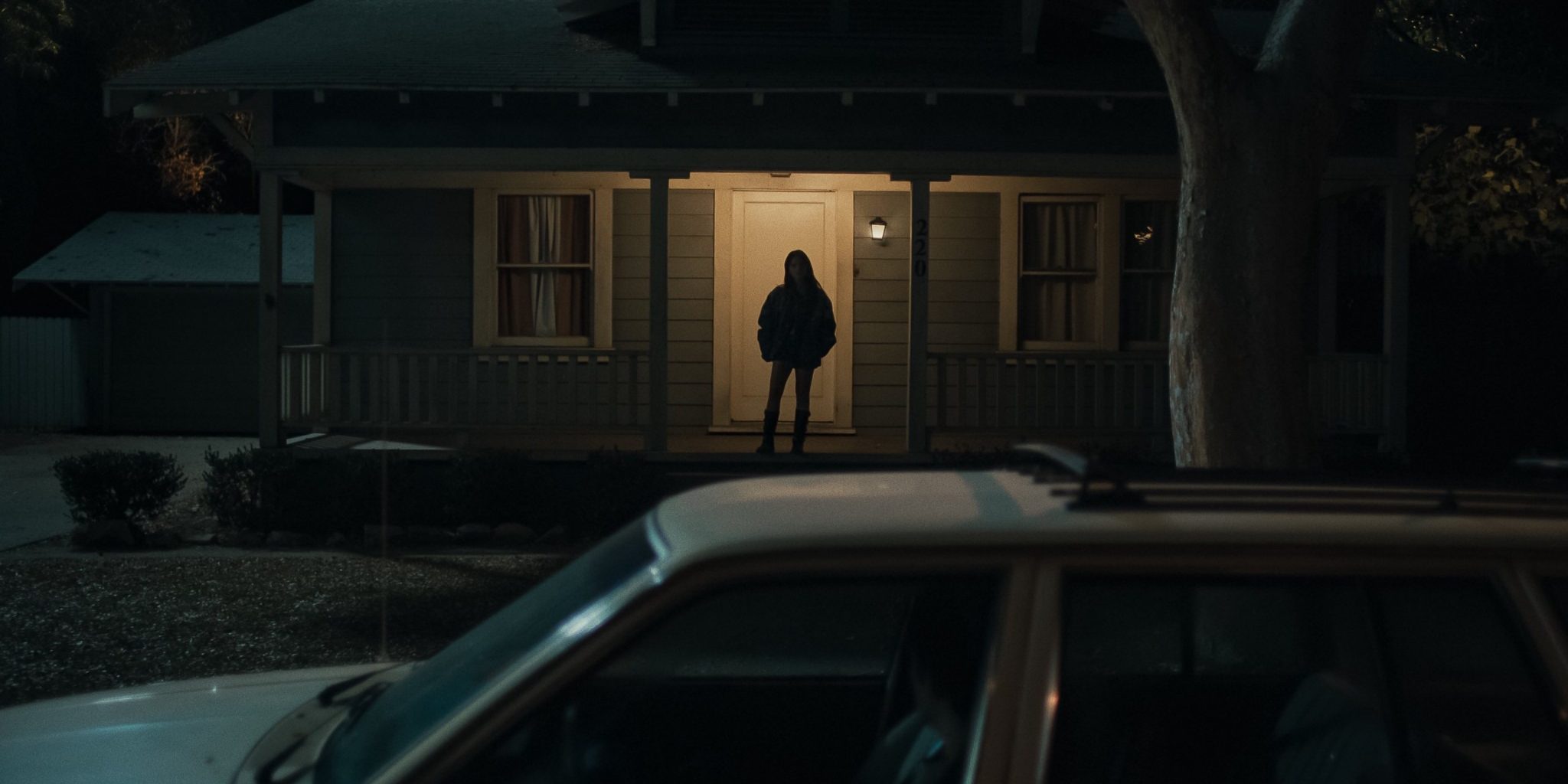 A car is parked outside of an American suburban home at night and a woman's silhouette stands in the porchlight