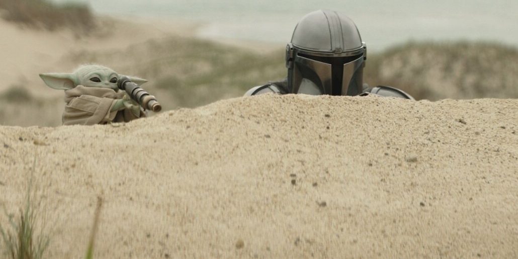 Mandalorian and Grogu peeking over a sandy ridge on a beach